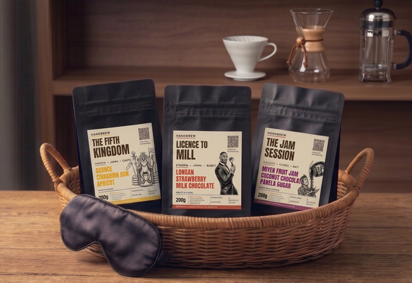 Three coffee packages in a basket with a sleep mask on a wooden surface, a v60, chemex, and a cafetiere in the background.