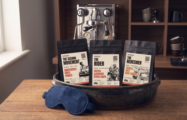 Three coffee bags labeled 'The Second Movement', 'The Rider', and 'The Redeemer' on a wooden table with a sleep mask and coffee-making equipment in the background.